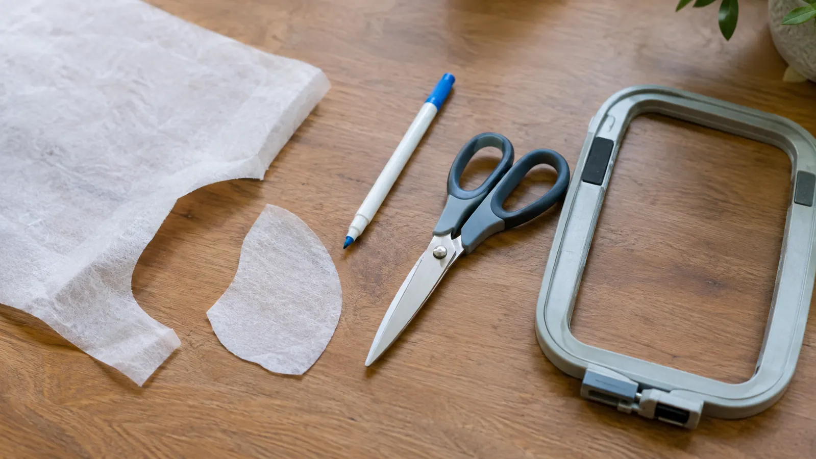 Embroidery prep supplies on a wooden work table including backing, scissors, and a magnetic hoop